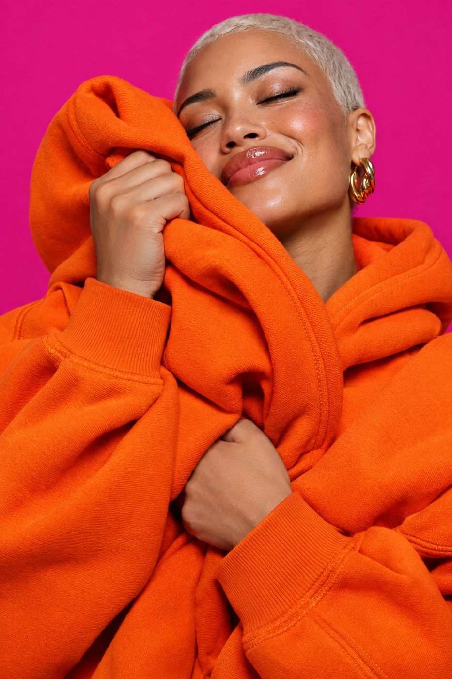 Creadora abrazando una sudadera naranja con los ojos cerrados sobre fondo magenta